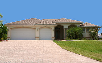 Front View A Generic Florida Home Sits On A Small Grass Covered Hill With Garage, Palm Trees And A Clear Blue Sky, Looking Up The Driveway.